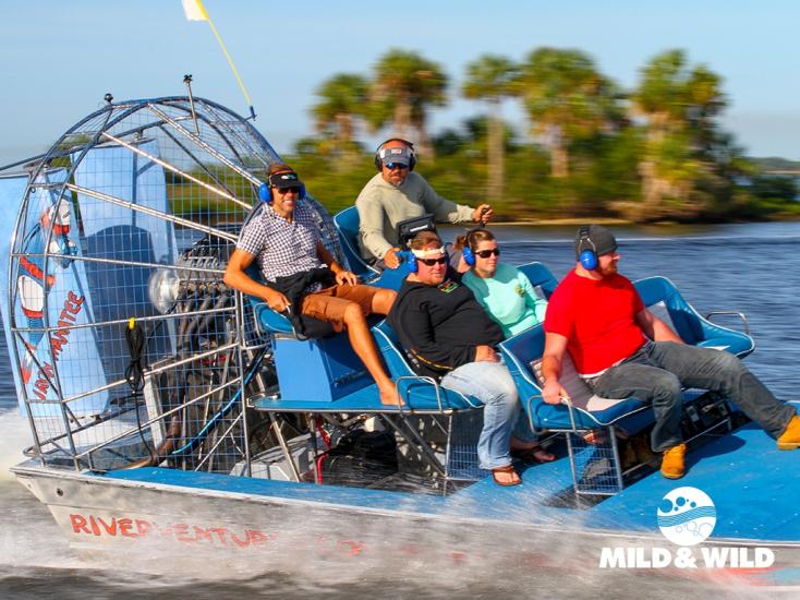 Four guests and a driver sit atop an airboat flying through water at a high speed while the land and water blurs around them.